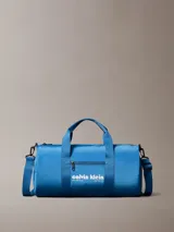 Cartera tipo duffle deportiva de color azul brillante, confeccionada en poliéster. Presenta asas de mano y una correa ajustable para el hombro. En el centro tiene estampado el logo de Calvin Klein en blanco, incluyendo la dirección "West 39th Street New York City". Posee un bolsillo exterior con cierre en el frente.