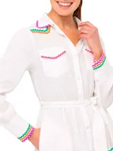 Vestido camisero largo de lino blanco con cuello camisero, cierre frontal con botones y cinturón de lazo. Presenta detalles de bordado ondulado en verde, naranja y rosa en cuello, bolsillos, puños y ruedo.
