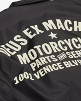 Campera negra tipo coach con cierre de botones a presión, bolsillos laterales y logo de la marca Deus Ex Machina estampado en el pecho.