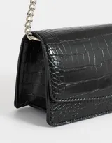 Bandolera negra de cuerina con textura croco, solapa con broche a presión y correa de cadena metálica plateada con charm lateral.