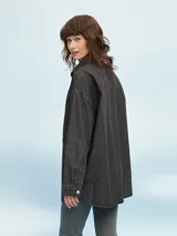 Camisa de denim negro con corte oversize, cuello clásico, cierre frontal con botones metálicos y bolsillo de parche en el pecho.