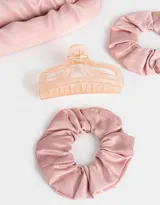 Set de accesorios para el cabello que incluye un rizador sin calor de tela satinada color rosa, dos scrunchies de tela satinada color rosa y un broche de plástico color durazno.