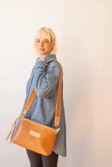 Cartera bandolera de cuero color marrón con textura croco. Tiene correa larga trenzada y cierre superior con borla decorativa.