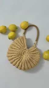 Bolso de mano tejido a mano con hilado de algodón grueso, de forma redonda y acanalada, color beige claro. Posee un asa trenzada de material similar al cuero.