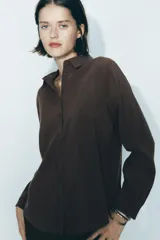 Camisa marrón de corte holgado, con cuello de solapa, manga larga con pliegues y bajo asimétrico. Cierre frontal con botones.