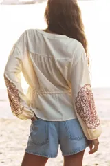 Blusa color crema de cuello redondo y manga larga con elástico en los puños. Presenta bordados florales en el frente y mangas, puntilla a tono y cintura ajustable con cordón y borlas. Cierre frontal con botones.