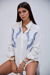 Camisa blanca de manga larga con cuello clásico y botones frontales. Presenta un diseño de ondas en tonos azules con lentejuelas y pequeños apliques metálicos en el frente.