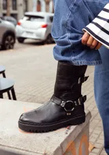 Bota de caña media color marrón oscuro, realizada en cuero encerado. Con hebillas en la caña y herrajes de níquel. El fondo está realizado artesanalmente, es de suela, y es negro. El interior está forrado en forrería vacuna.