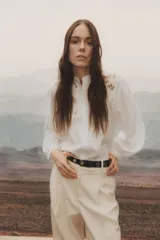 Blusa blanca de corte holgado en tejido liviano de lino y viscosa con canesú delantero en broderie. Cuello mao con frunces en la nuca, botones forrados adelante y mangas globo largas con puños finos abotonados. Bajo redondeado.