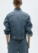 Chaqueta de jean azul de corte recto con cuello camisero, cierre frontal con botones metálicos, dos bolsillos con solapa en el pecho y mangas largas con puños abotonados.