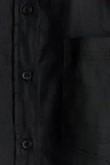 Camisa negra de lino liviano, con cuello clásico, cierre frontal con botones, canesú de doble capa en la espalda, hombros ligeramente caídos, mangas largas con puños abotonados, bolsillo de parche en el pecho y bajo redondeado.