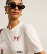 Remera blanca de algodón con cuello redondo y mangas cortas abullonadas. Presenta estampados de un lazo, una flor, un corazón y frases caligráficas.