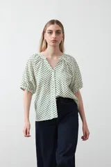 Camisa de viscosa con estampado de pequeños lunares verdes sobre fondo blanco o crema. Presenta escote en V con botones frontales y mangas cortas abullonadas con elástico en el puño.