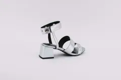 Sandalias plateadas con dos tiras horizontales sobre el pie y una tira vertical que se une a una pulsera con hebilla en el tobillo. La pulsera tiene ojales y tachas combinadas. El taco es cuadrado y de 5 cm.