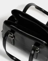 Cartera tipo city de cuerina color negro con efecto charol. Tiene manijas superiores con apliques metálicos en forma de corazón, cierre superior y bolsillos laterales con trenzado decorativo.