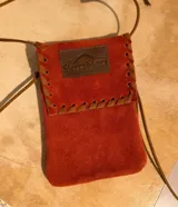 Cartera pequeña de gamuza color naranja con correa larga de cuero marrón para colgar al cuello. Tiene un aplique de cuero marrón con el logo de la marca Sierra Mora.