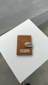 Porta pasaporte de cuero en color tostado con cierre metálico.