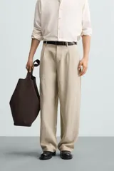 Pantalón de corte holgado (wide fit) en color beige, confeccionado en tejido con mezcla de lyocell. Presenta pliegues frontales en la cintura, bolsillos laterales y bolsillos traseros de doble vivo. Cuenta con cierre frontal de cremallera y botón.