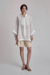 Camisa blanca de lino con cuello clásico, cierre frontal con botones y mangas anchas 3/4. Presenta detalles de bordado calado en el frente y puños.
