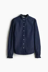 Camisa de denim azul oscuro con cuello mao y volados en cuello y puños. Tiene mangas largas abullonadas con botón en los puños, botonadura frontal y bajo redondeado.