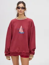 Buzo color borgoña de corte clásico con cuello redondo y puños acanalados a rayas. Estampado frontal de un velero.