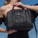 Cartera tipo baúl color negro, de cuero sintético, con doble asa corta y correa larga regulable y desmontable. Tiene cierre superior, dos compartimentos internos y tres bolsillos internos, uno con cierre.