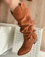 Bota tejana de caña alta color marrón tostado, confeccionada en cuero vacuno con detalle de flecos en el lateral exterior. Tiene taco de 4 cm y puntera fina. La caña puede usarse alta o arrugada.