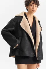 Chaqueta corta de poliéster con efecto cuero en color negro e interior de corderito beige. Presenta un diseño de corte holgado con solapas anchas, cierre cruzado y bolsillos laterales.