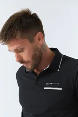 Polo de manga larga negra con cuello clásico, tapeta de dos botones y bolsillo de parche en el pecho con ribete blanco. Logo de la marca bordado en blanco sobre el bolsillo.