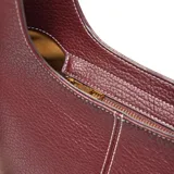 Bolso tipo hobo de cuero sintético (PU) color bordó, con costuras blancas visibles en el centro. Posee un asa corta fija y una correa larga desmontable y regulable con tejido a rayas blancas y negras. Cuenta con cierre metálico superior y herrajes dorados.