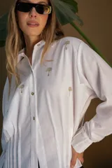 Camisa blanca de lino con bordados dorados de árboles, corte recto, cuello camisero y mangas largas con puños abotonados.