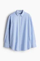 Camisa Oxford de algodón a rayas verticales celestes y blancas, con cuello clásico, botones frontales, canesú en la espalda, corte relajado, hombros caídos, mangas largas con puños sencillos y bajo redondeado con espalda más larga.