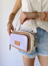 Bandolera rectangular de cuero color lila, con detalles en marrón y blanco. Tiene correa larga regulable, bolsillo frontal con cierre y etiqueta de cuero con logo de la marca.