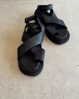 Sandalias bajas color gris topo, con tiras anchas cruzadas sobre el empeine y tobillera con hebilla metálica. Suela plana de goma negra.