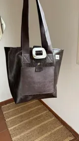 Bolso tote de ecocuero marrón oscuro con textura cocodrilo y un parche frontal de cuero liso marrón claro. Posee asas de hombro de cuero marrón y un broche decorativo blanco en el frente.