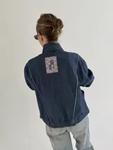 Campera de jean negra tipo bomber con cuello redondo y cierre frontal con botones metálicos. Presenta un parche rectangular con diseño gráfico en la espalda.