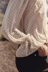 Blusa color crema semitransparente con cuello redondo con volantes y cordón. Manga larga acabada en elástico y volante. Cierre frontal con botones.