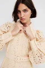 Vestido corto color beige con broderie, cuello camisero, botonadura frontal, cinturón extraíble con hebilla redonda, mangas globo largas con puños abotonados y ribetes festoneados.