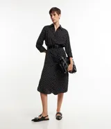 Vestido midi tipo chemise de tela satinada negra con lunares blancos, cuello solapa, abotonado delantero, mangas largas y cinturón con hebilla.