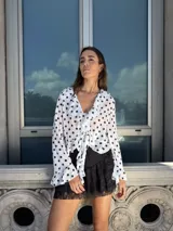 Blusa blanca traslúcida con estampado de lunares negros, escote en V con volados y lazo en la cintura, mangas largas acampanadas.
