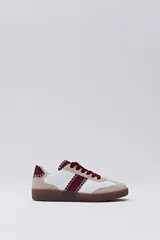 Zapatilla deportiva de estilo retro, con base blanca, puntera y talón en gamuza beige, y franja lateral y cordones en color bordó. Presenta costuras decorativas en contraste en los bordes de las piezas de gamuza.