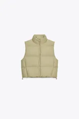 Chaleco blanco acolchado con cuello alto y cierre frontal con cremallera. Tiene bolsillos delanteros con cierre de cremallera oculta y bajo ajustable con cordones elásticos laterales.