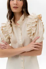 Camisa de lino color lila con volados en el cuello y en las mangas cortas abullonadas. Presenta un diseño de corte recto con cierre frontal de botones.