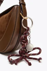 Accesorio charm para bolso con cuerda anudada en tonos borgoña, azul y beige, con abalorios metálicos plateados.