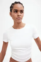 Remera blanca de corte cropped, con cuello redondo y mangas cortas. Presenta costuras fruncidas en un hombro y a ambos lados de la cintura, creando un efecto drapeado.