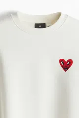 Buzo blanco de corte holgado con cuello redondo y puños y bajo acanalados. Presenta un pequeño logo de corazón rojo con ojos en el pecho.