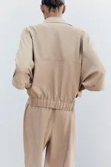 Camisa color beige de cuello solapa y manga corta con trabilla y botón. Tiene bolsillos delanteros de plastrón con solapa, bajo con elástico y cierre frontal con botones a presión.