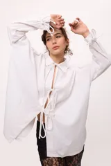 Camisa blanca de lino y algodón, con cuello clásico, cierre frontal con lazos y mangas largas abullonadas con puños ajustables con lazos.