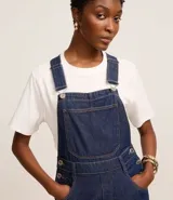 Jardinero largo de jeans azul oscuro, con breteles anchos ajustables con botones, bolsillo tipo parche en el pecho y cierre lateral con botones.