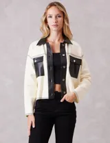 Chaqueta de corte boxy en color crema, confeccionada en tejido de punto suave con detalles en cuero sintético negro en cuello, solapas, cartera frontal y bolsillos. Presenta cierre frontal con botones metálicos plateados, hombros caídos y costuras decorativas horizontales.